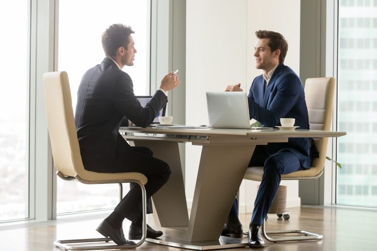 Deux hommes en costume échangent autour d'une table de réunion, devant une grande baie vitrée