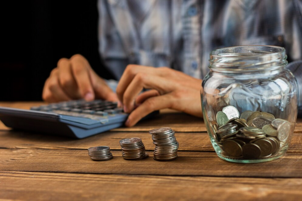 Mains sur une calculatrice, à côté de piles de pièces et d'un bocal d'économies